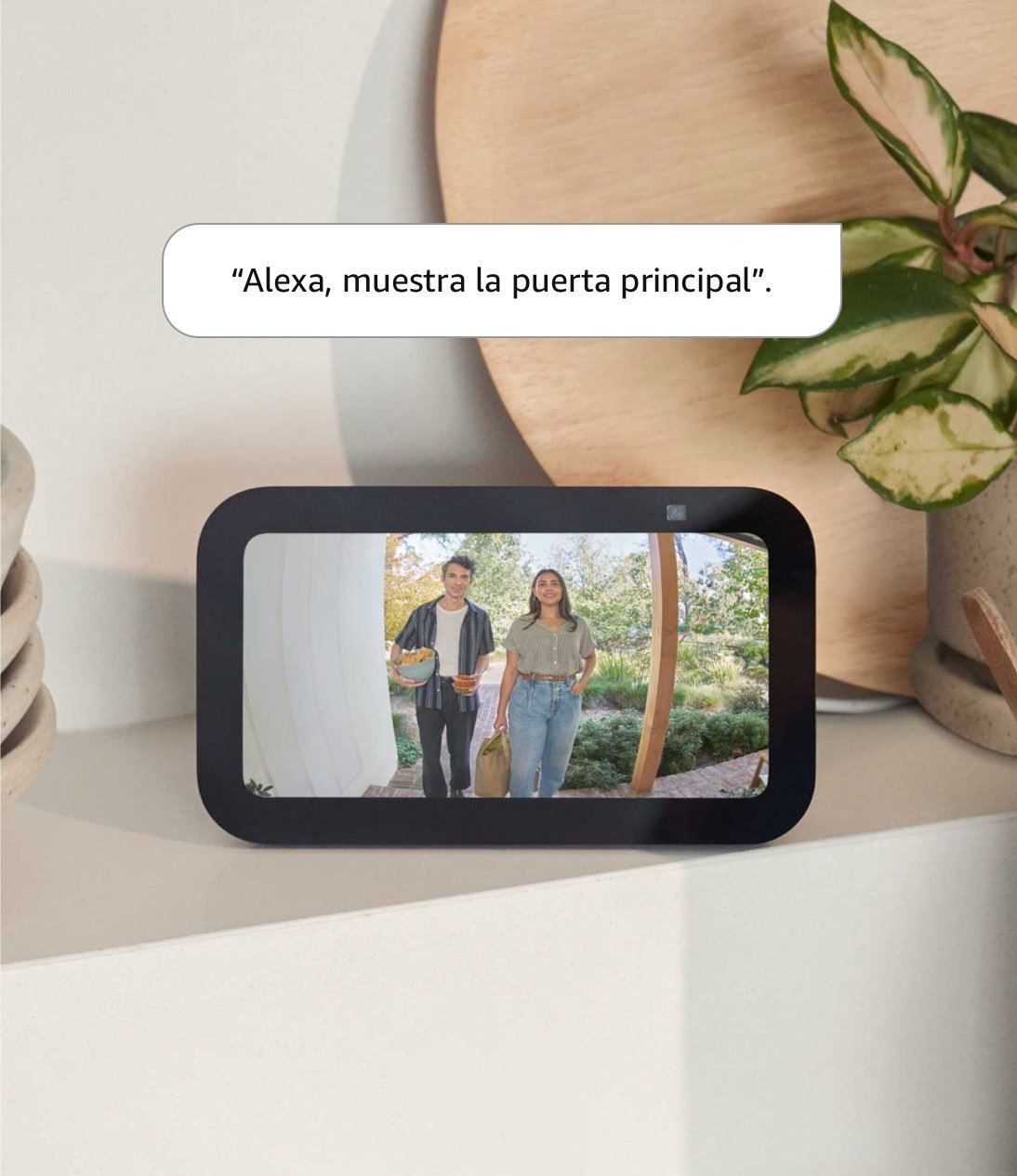 Echo Show 5: Una tablet de 5.8 x 3.4 in está en un mueble en posición horizontal. La pantalla muestra a un hombre y una mujer con botanas en un porche frontal. El texto dice “Alexa, muestra la puerta principal”.