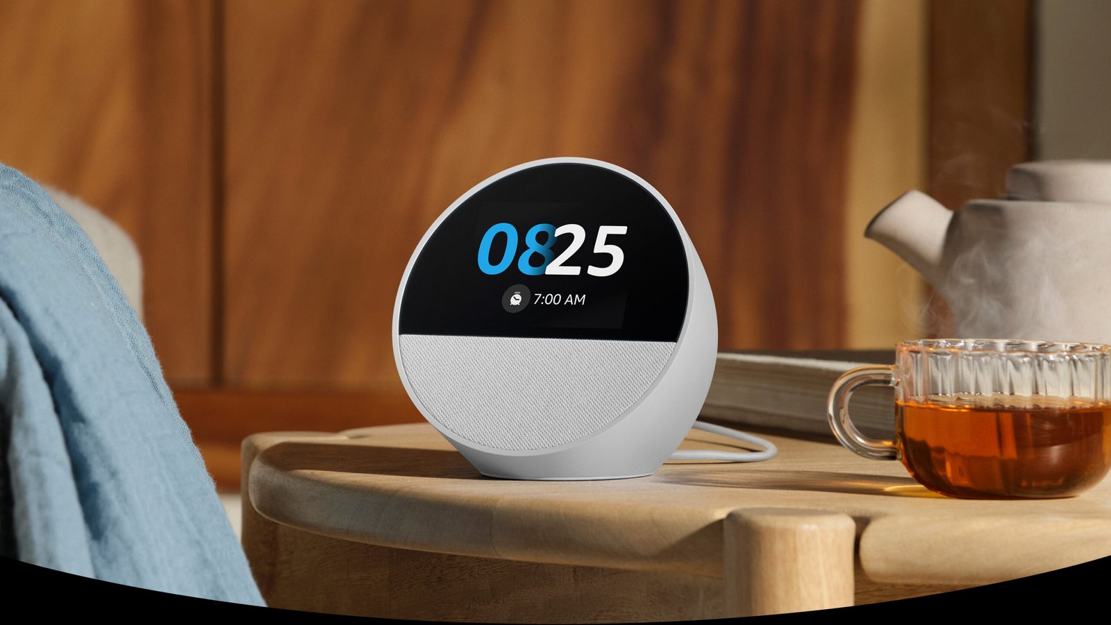 El Echo Spot, un parlante inteligente semiesférico con pantalla táctil de 2.8 pulgadas y parlante de malla en la parte inferior, está sobre una mesita de noche. La pantalla muestra la hora, la fecha, el clima y un ícono de clima soleado.