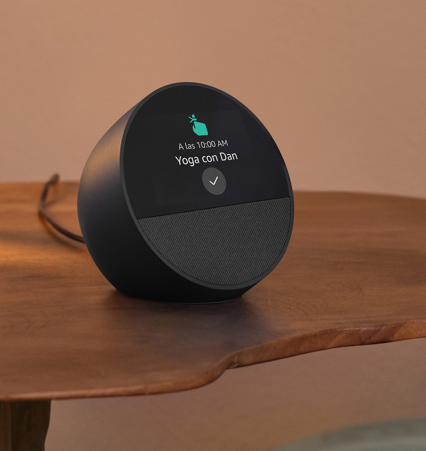 El Echo Spot está sobre una mesita de noche. La pantalla de 2.8 pulgadas muestra el recordatorio “Yoga con Dan a las 10 de la mañana” y un ícono de recordatorio con un dedo apuntando al dispositivo.