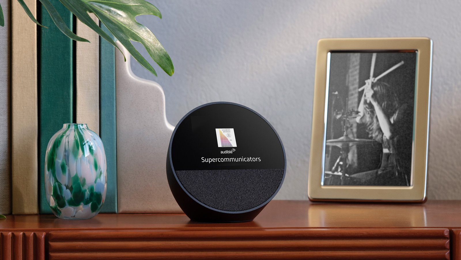 El Echo Spot está en una mesa de entrada al lado de una foto enmarcada y un conjunto de libros. La pantalla de 2.8 pulgadas muestra la hora y la portada del libro Supercomunicators en Audible.
