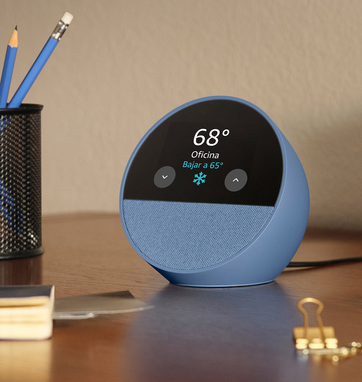 El Echo Spot está en un escritorio. La pantalla de 2.8 pulgadas muestra el termostato de la oficina en casa y el clima. El ícono de termostato incluye el texto “Bajando a 65 grados”.